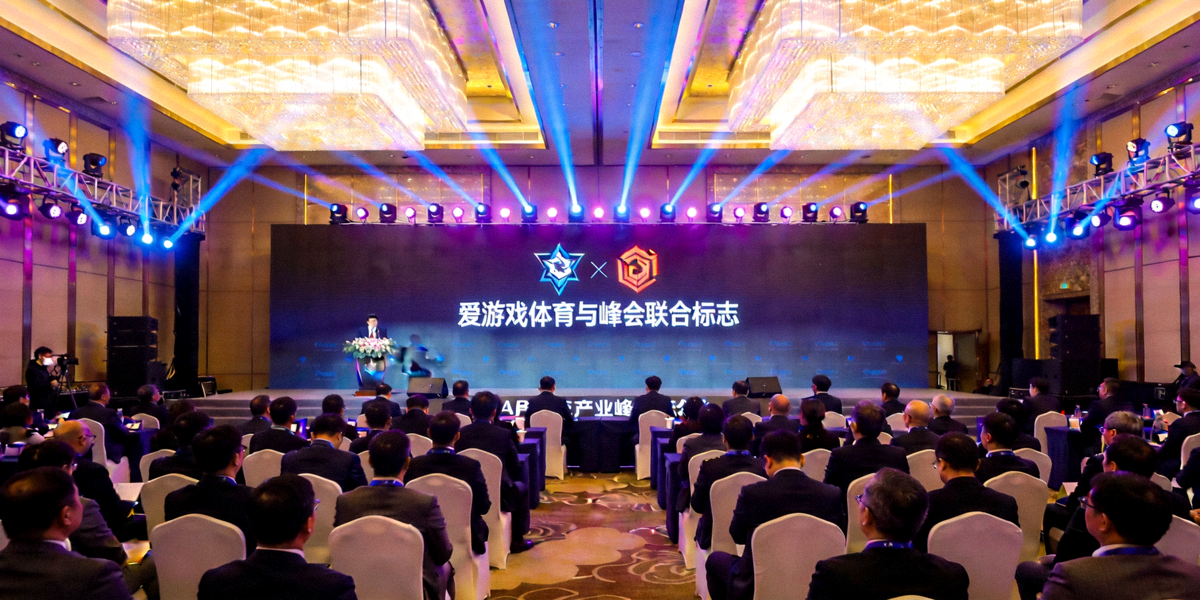 电竞产业峰会高峰论坛主会场全景，舞台灯光璀璨，大屏幕显示爱游戏体育与峰会联合标志，台下坐满行业精英